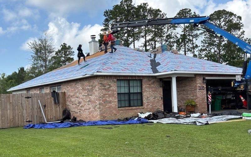 Crew on the roof of a brick house during a tear-off, with a blue boom lift and tarps on the lawn